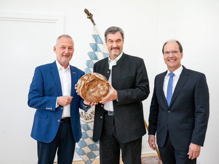 Bild von Landesinnungsmeister Günther Wagner und Geschäftsführer Stephan Kopp und Ministerpräsident Dr. Markus Söder, alle stehen vor einer bayrischen Flagge und Herr Söder hält ein Leib Brot in den Händen 